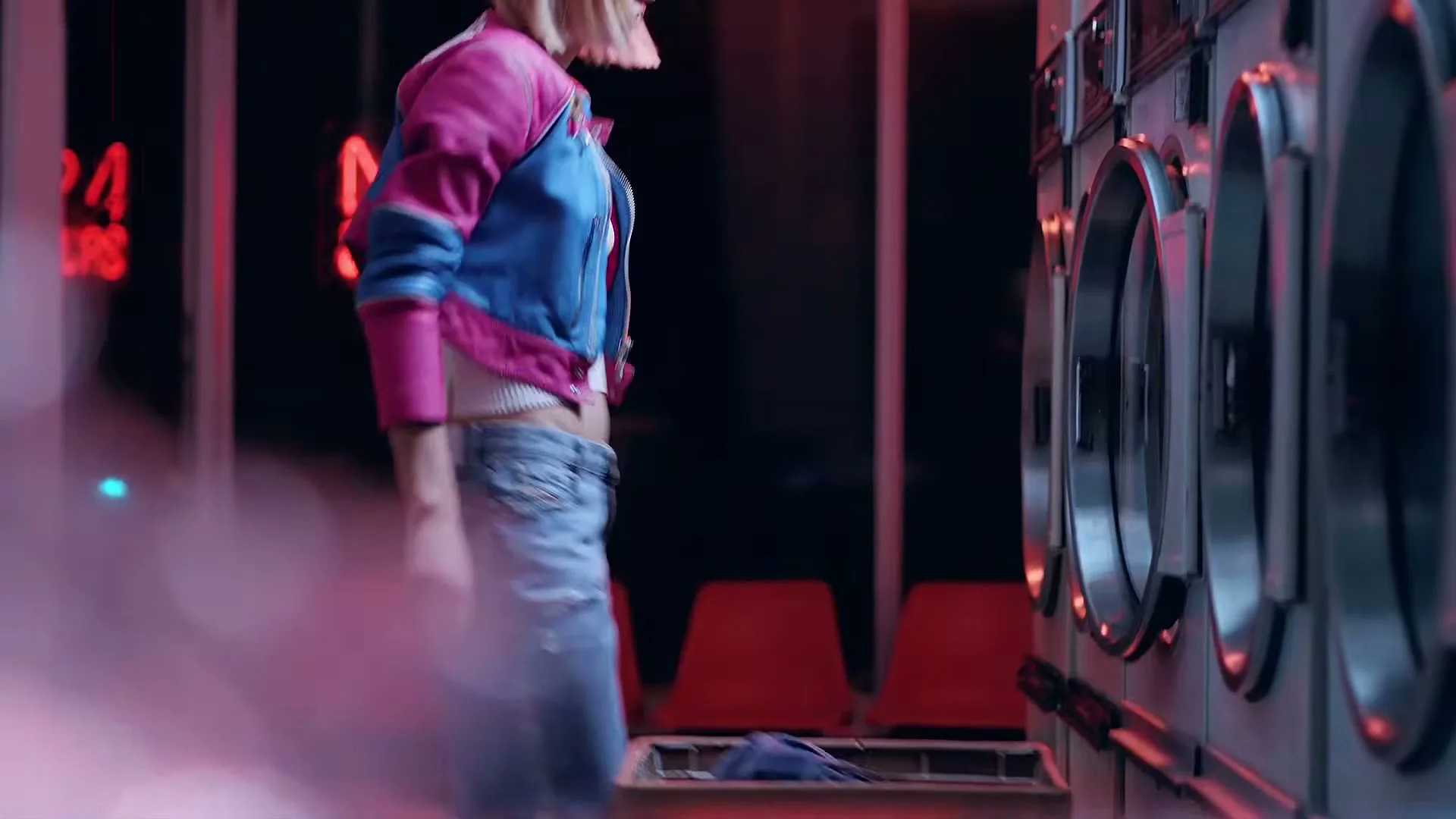 A young woman with blonde hair, wearing a pink and blue jacket and jeans, bends over a laundry basket in a neon-lit laundromat.