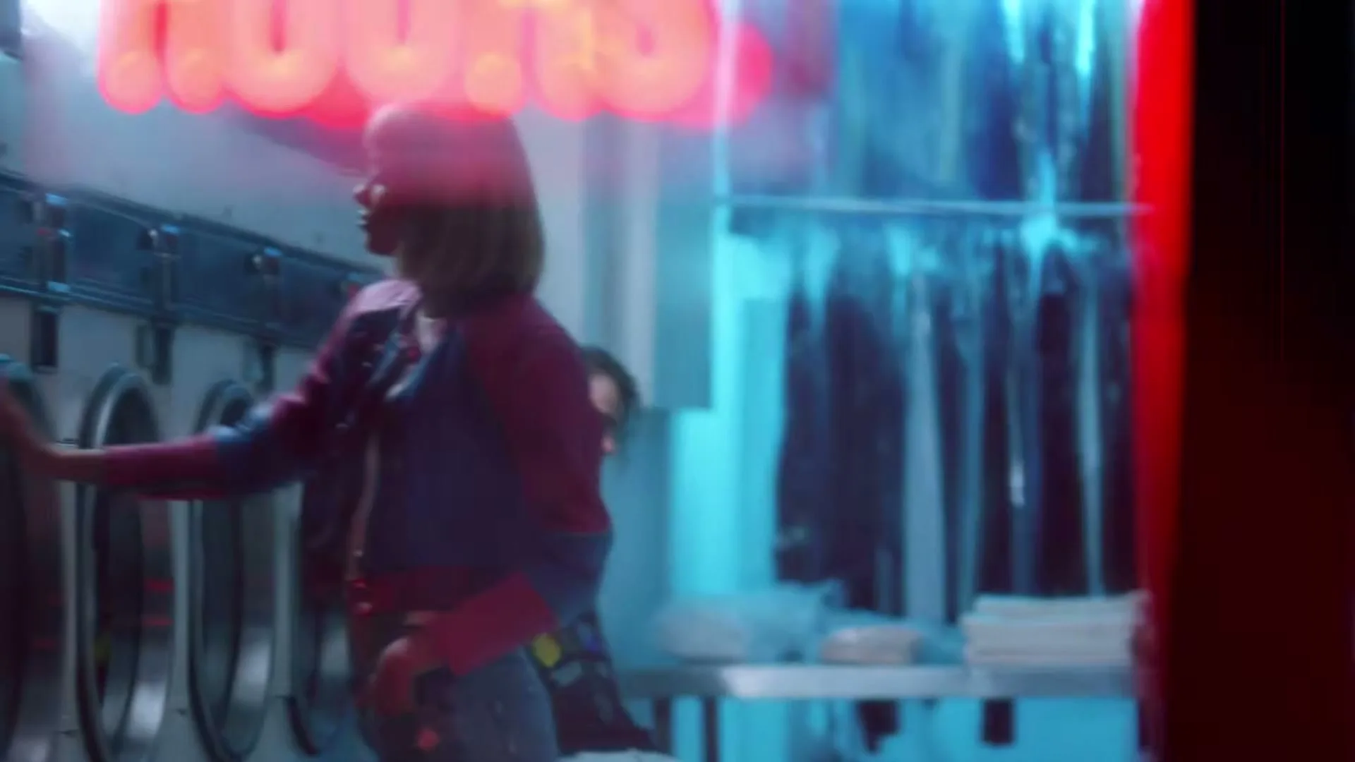 A young woman with a bob haircut, wearing a blue and red jacket and jeans, loads laundry into a washing machine in a laundromat at night.