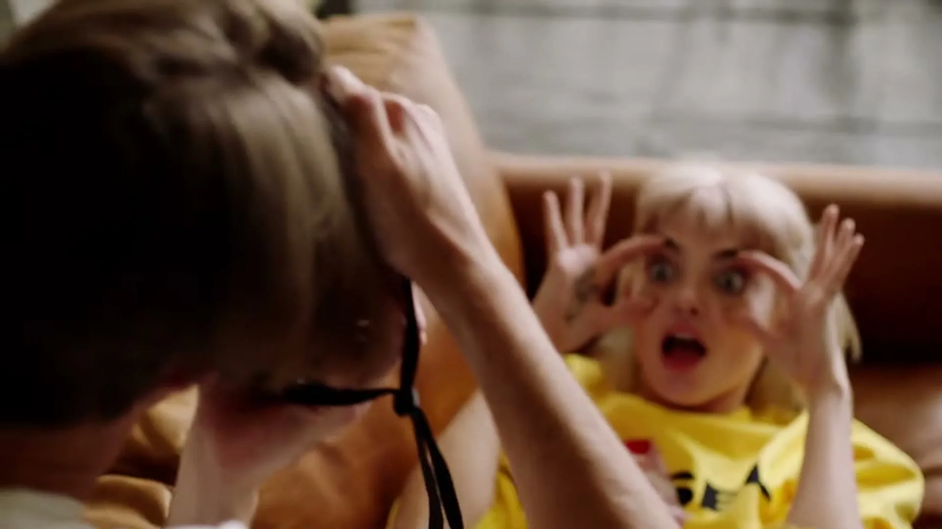 A blonde woman in a yellow sweatshirt lies on a brown couch, making a silly face with her eyes rolled back and tongue out, while a person with brown hair adjusts something in the foreground.