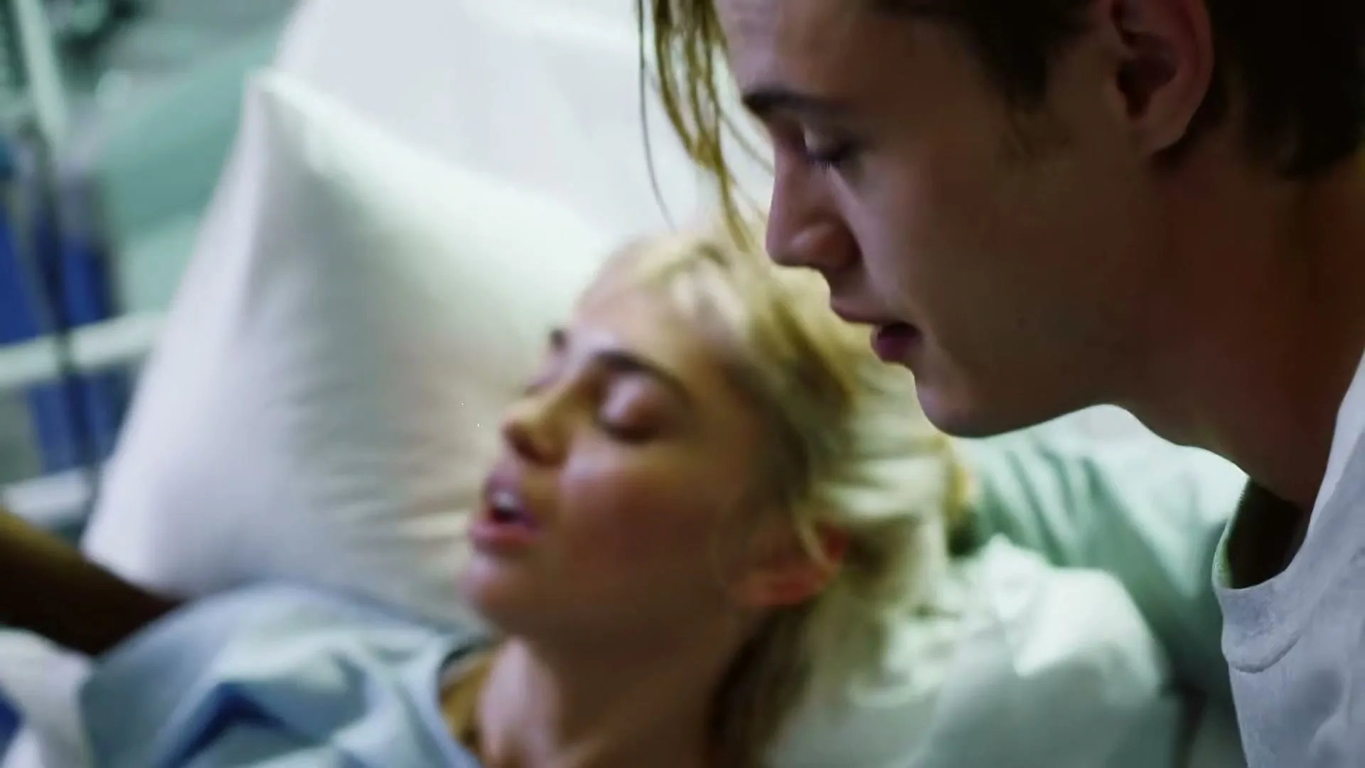 A young man with long hair looks down at a young woman with blonde hair, who is lying in a hospital bed, appearing to be in pain.