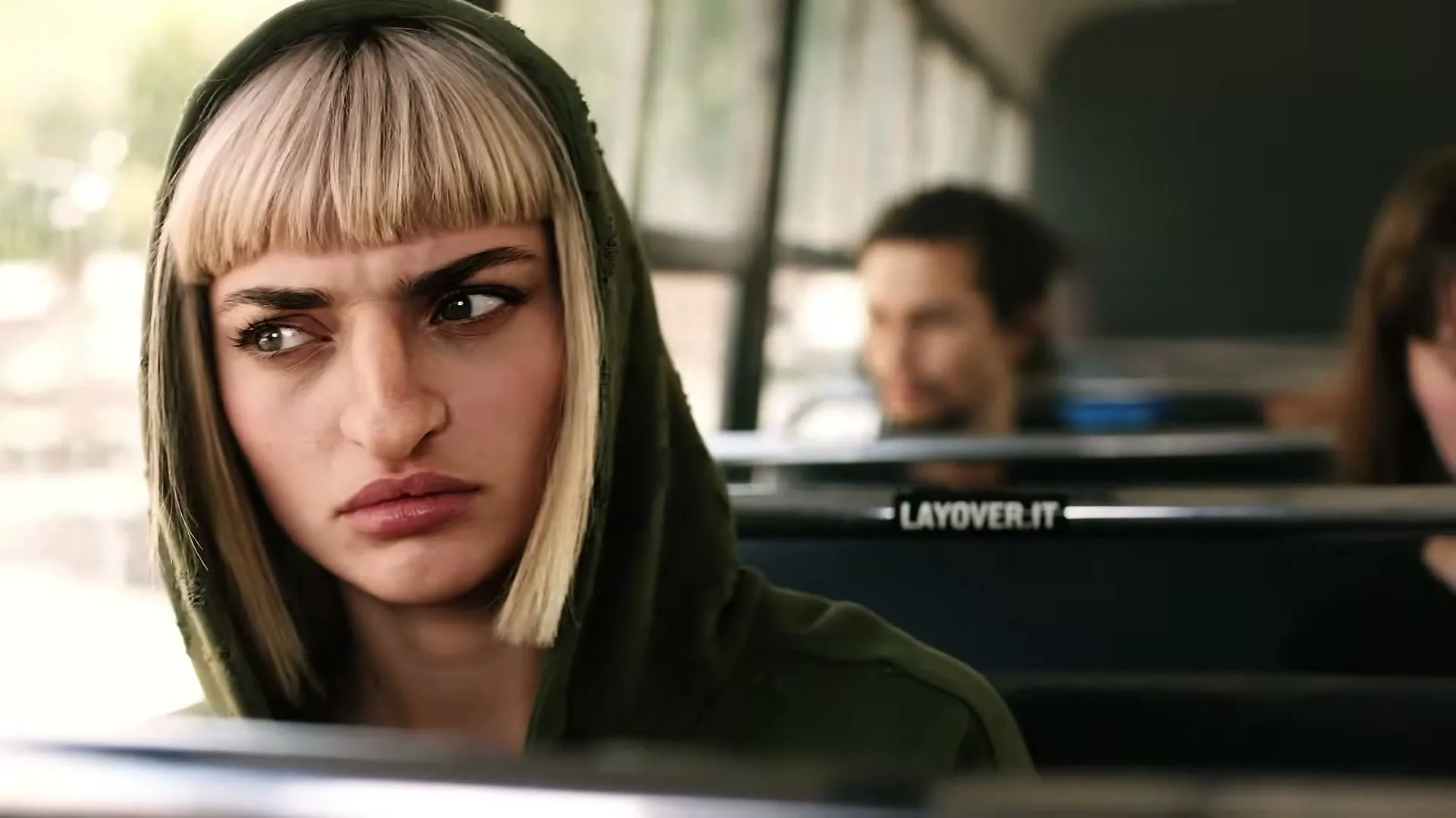 A young woman with blonde hair and a green hoodie sits on a bus, looking around with a concerned expression.