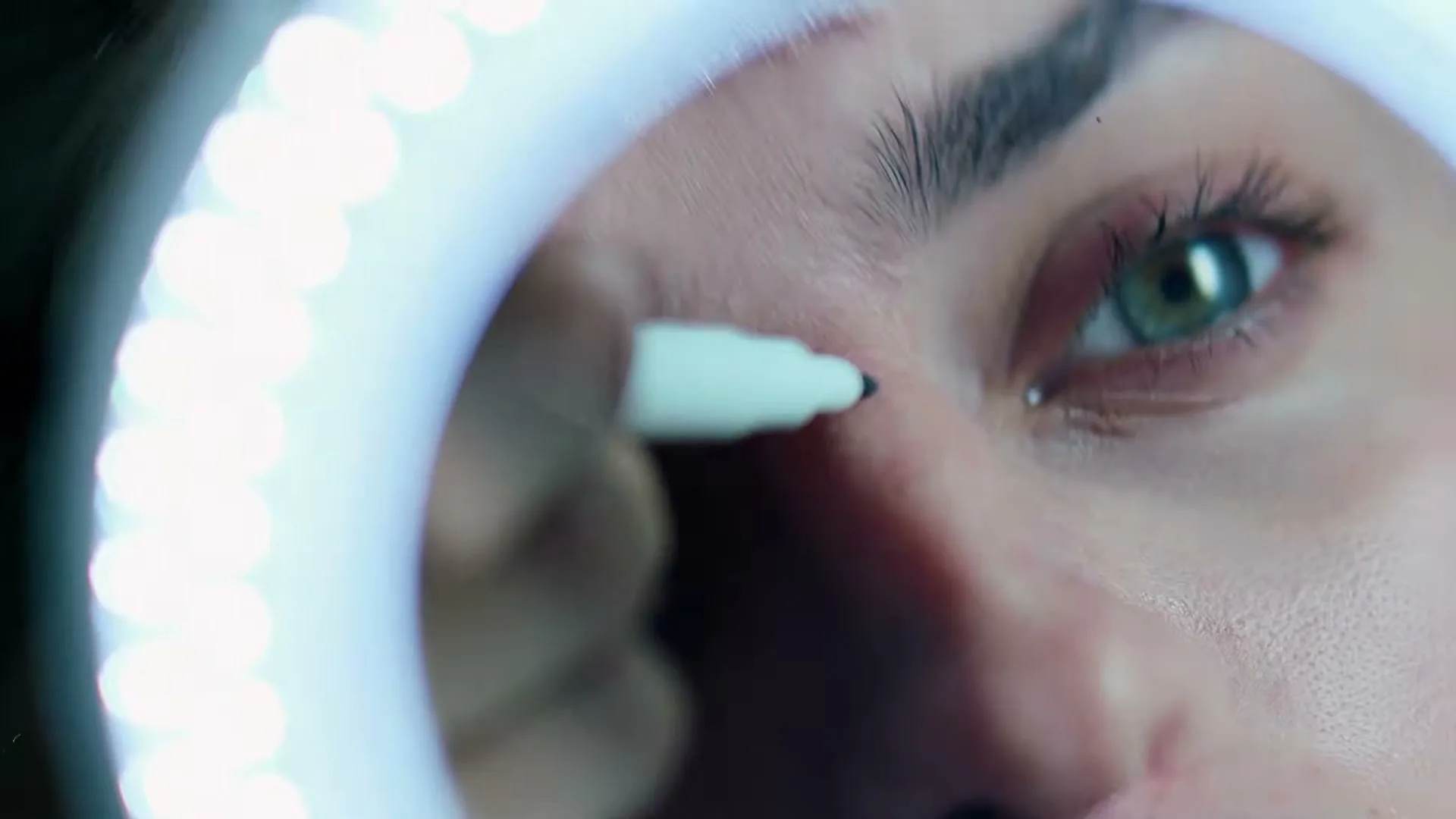 A woman with green eyes uses a white marker to draw on her face, framed by a glowing ring light.