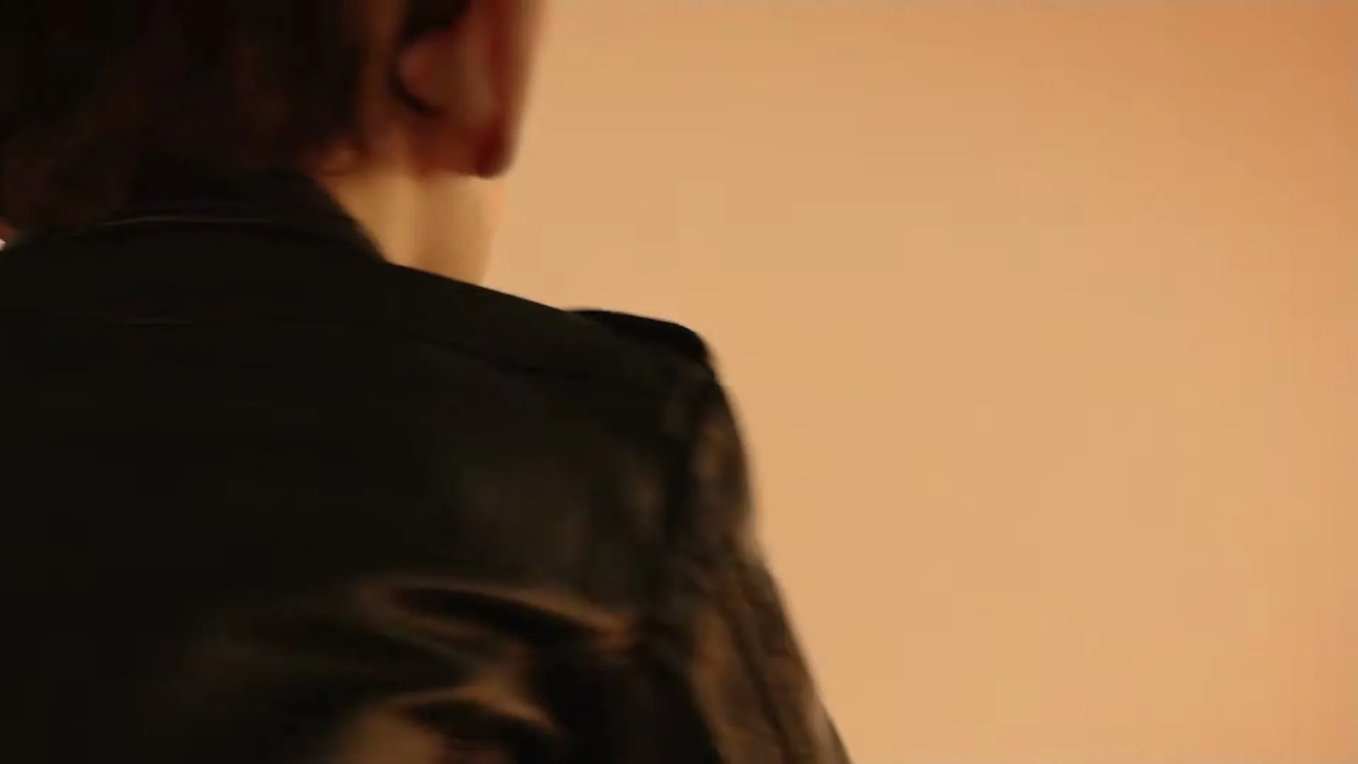 A young boy with large ears and a black leather jacket laughs joyfully, looking up against a warm, plain background.