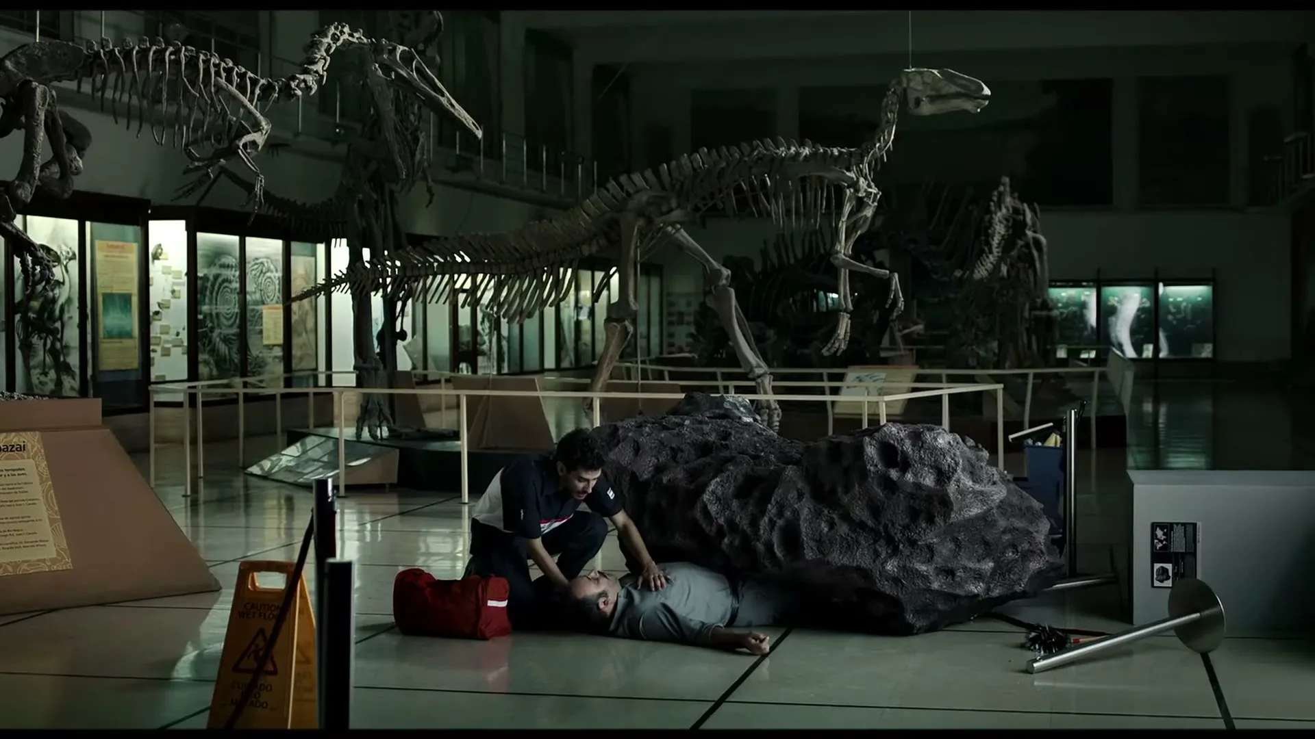 A man in a dark blue shirt and black pants performs CPR on another man lying on the floor, partially covered by a large, dark, rock-like object, in a dimly lit museum with dinosaur skeletons in the background.