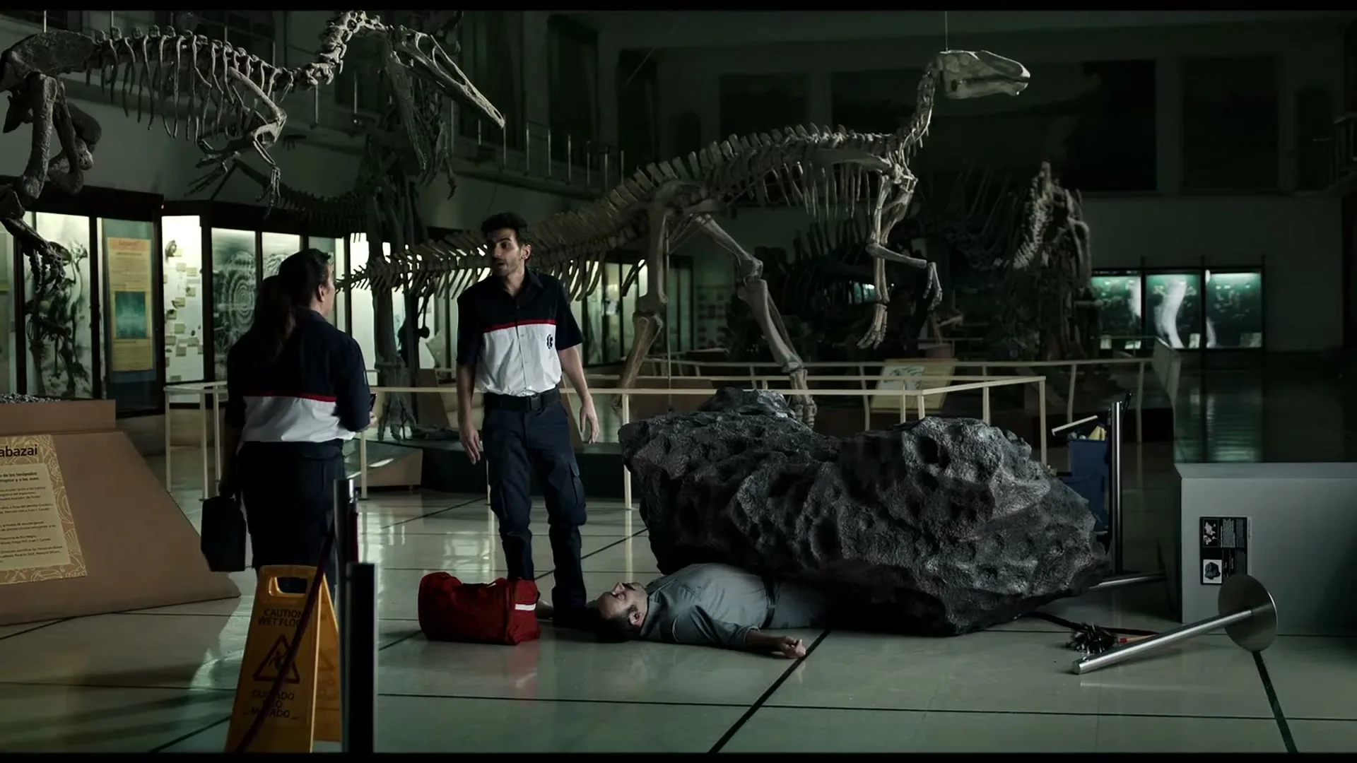 Two museum workers, a man and a woman, stand over a third man lying on the floor, partially covered by a large rock, in a dimly lit natural history museum.