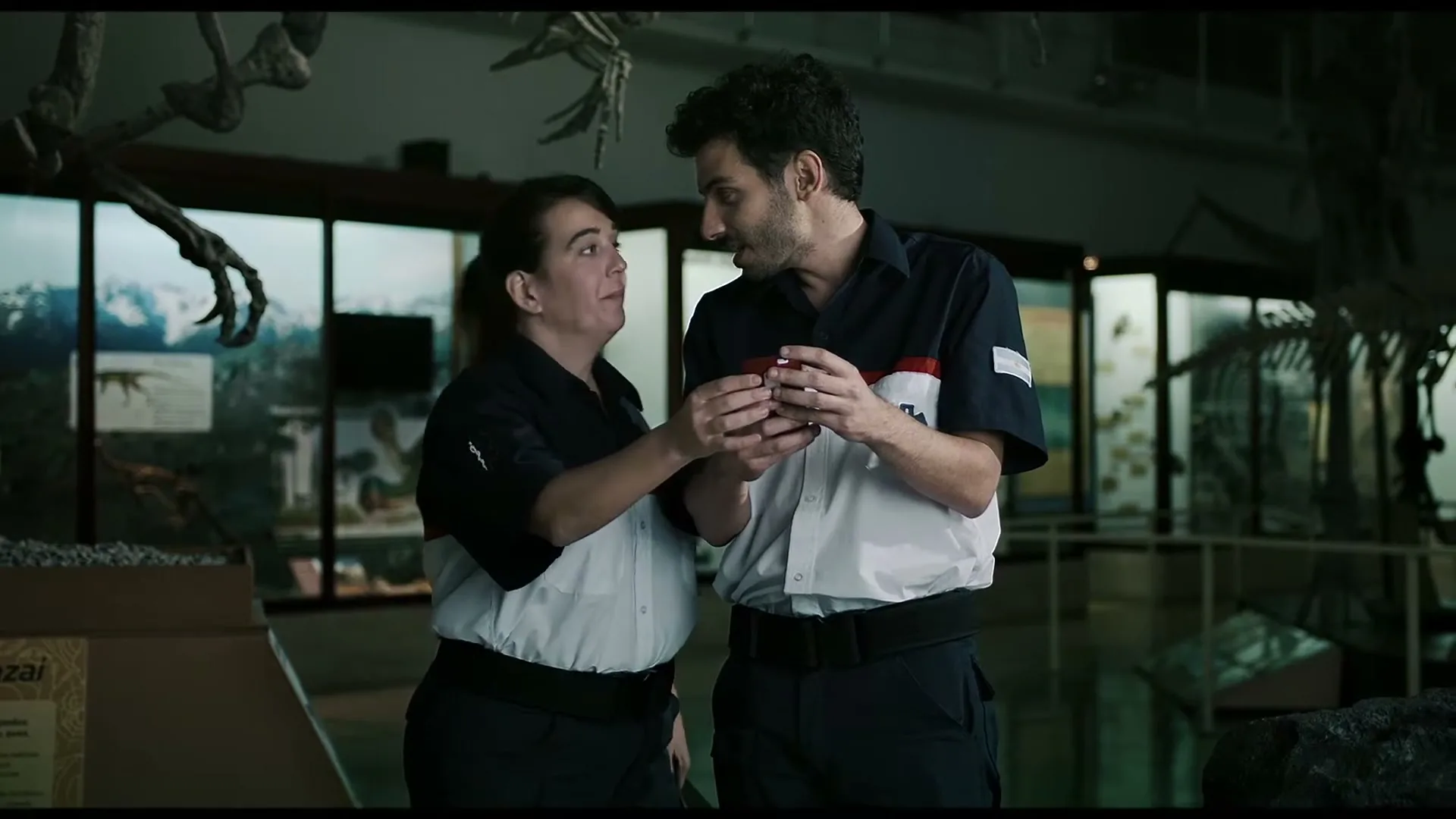 Two museum workers, a man and a woman, in uniform, look at a small red object in the man's hand, smiling and talking in a museum.