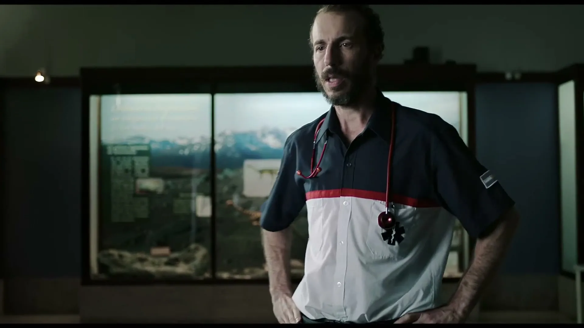 A male paramedic with a beard and stethoscope around his neck stands in a museum, looking around with a curious expression.