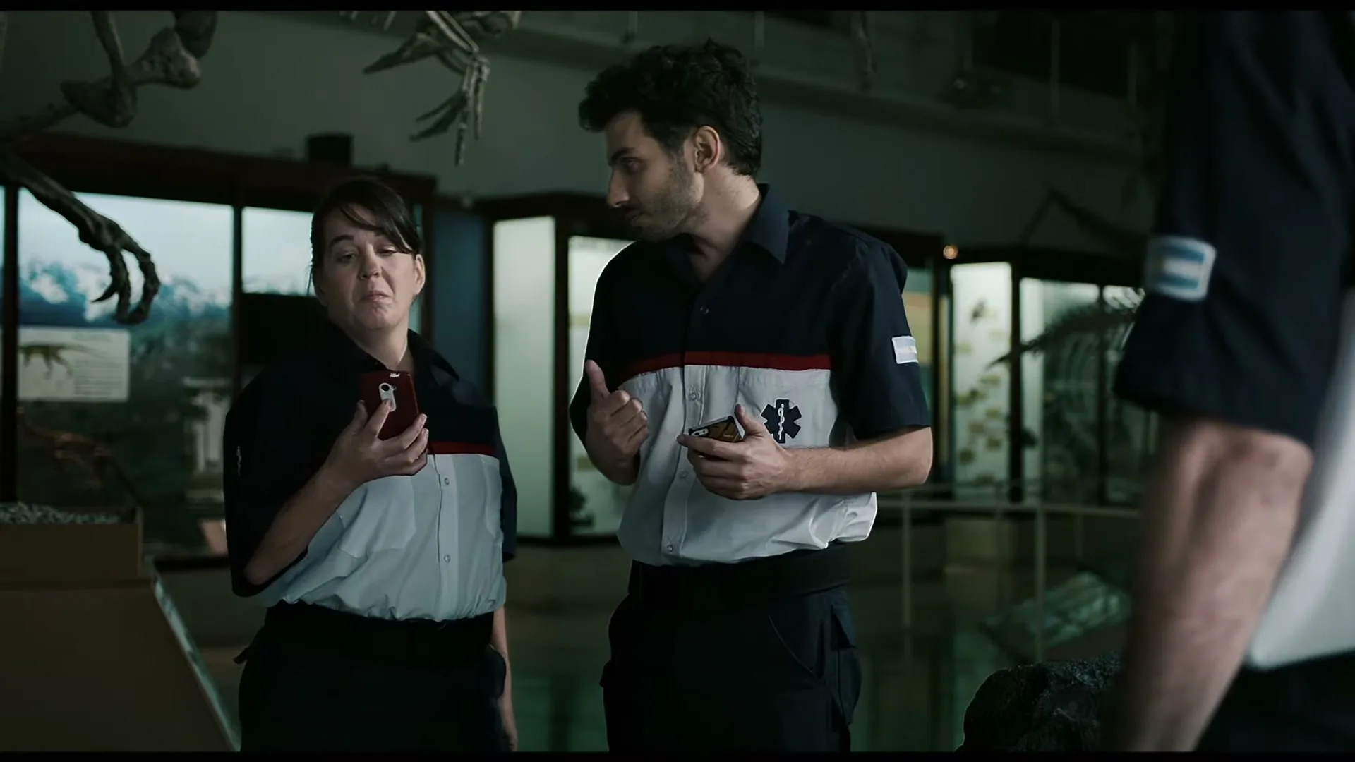 Two paramedics, a man and a woman, stand in a museum, looking up and talking to someone off-screen, holding their phones.