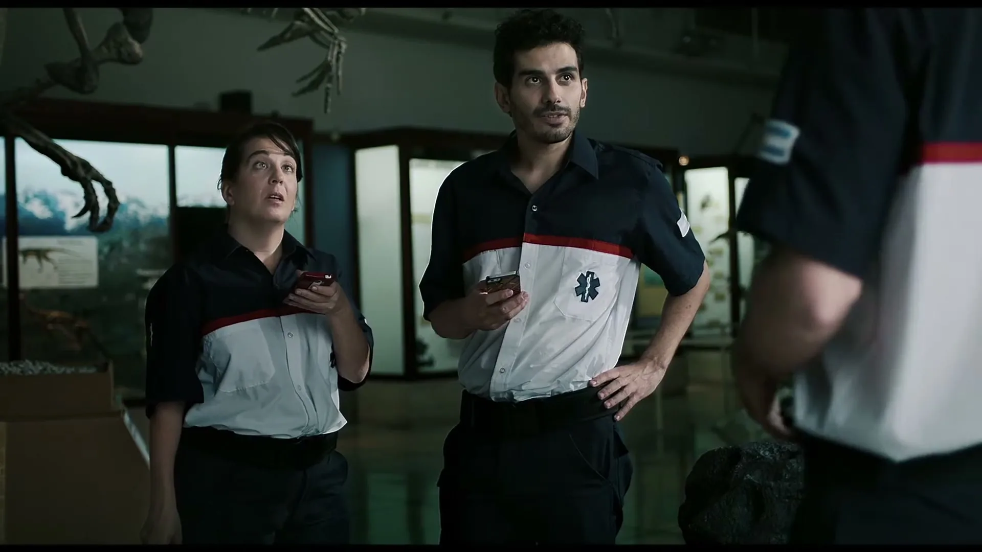 Two paramedics, a woman and a man, stand in a dimly lit museum, looking up and holding phones.
