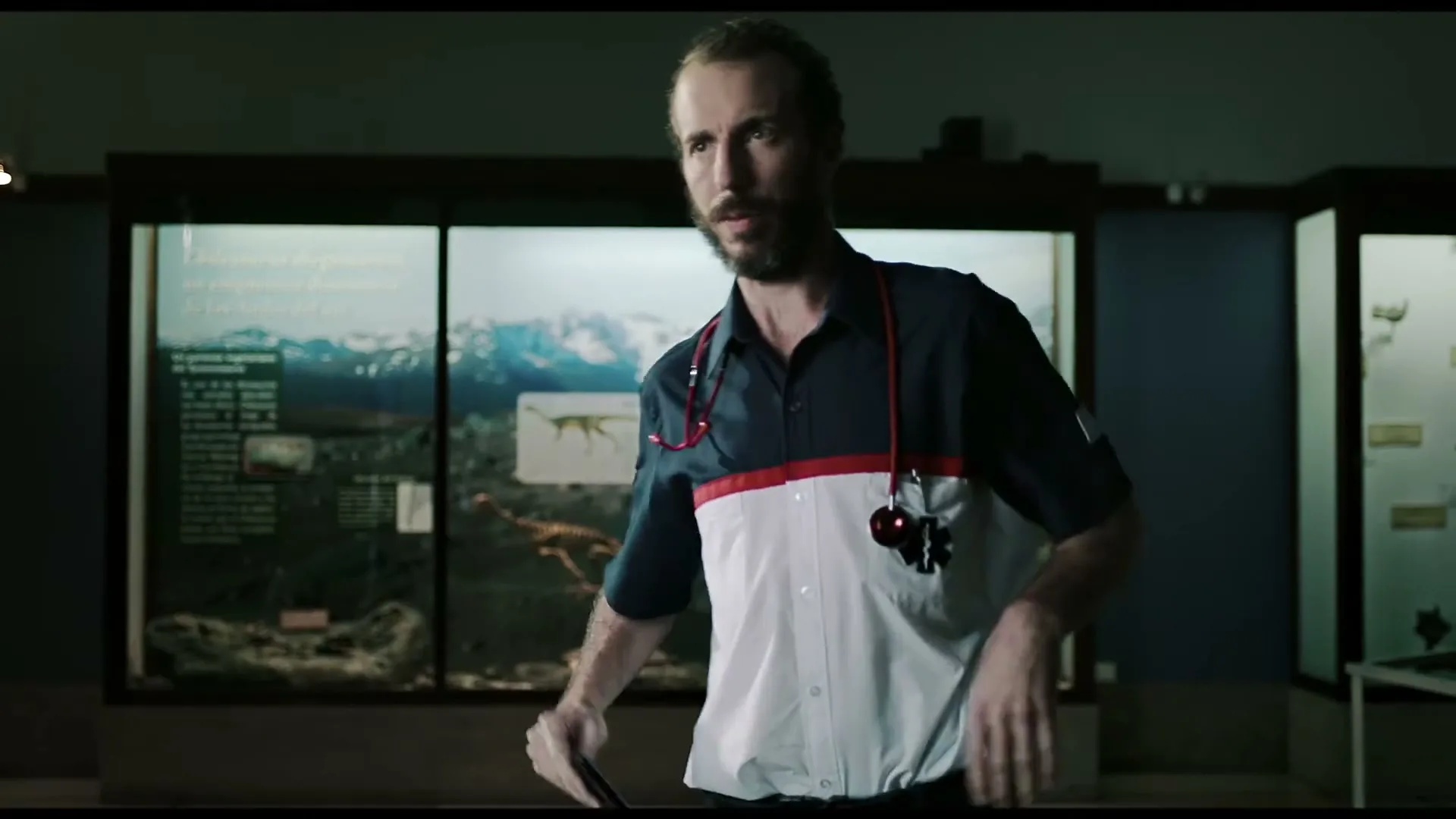 A male museum employee with a beard and a two-toned uniform shirt holds a smartphone in a dimly lit museum with display cases.