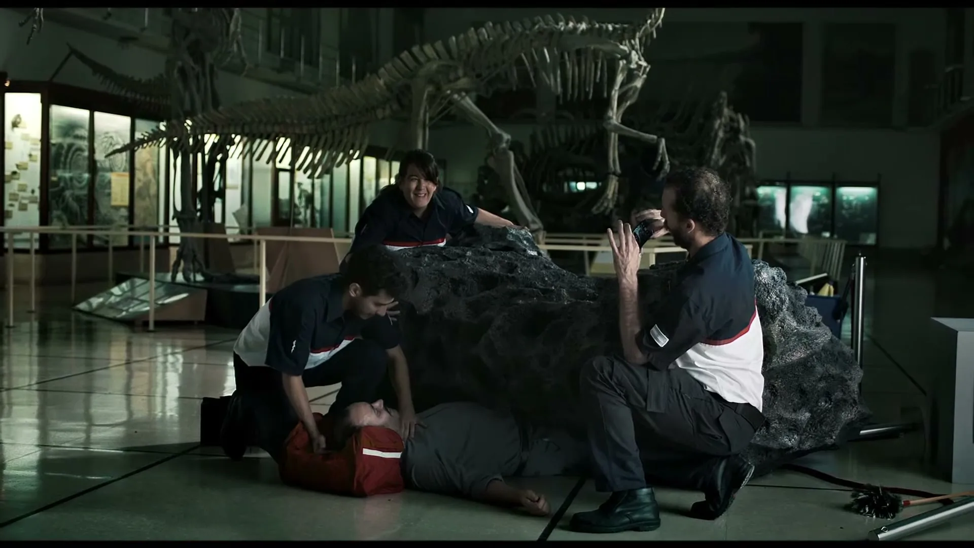 Three paramedics in uniform are gathered around a man lying on the floor, pinned under a large meteorite, inside a museum with dinosaur skeletons.
