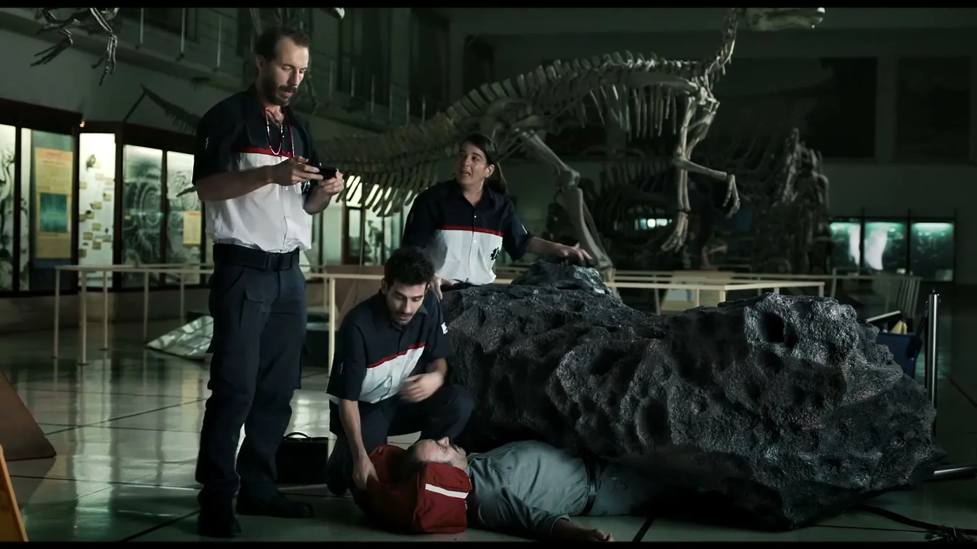 Three museum workers in uniform attend to a man lying on the floor next to a large rock, with dinosaur skeletons in the background.