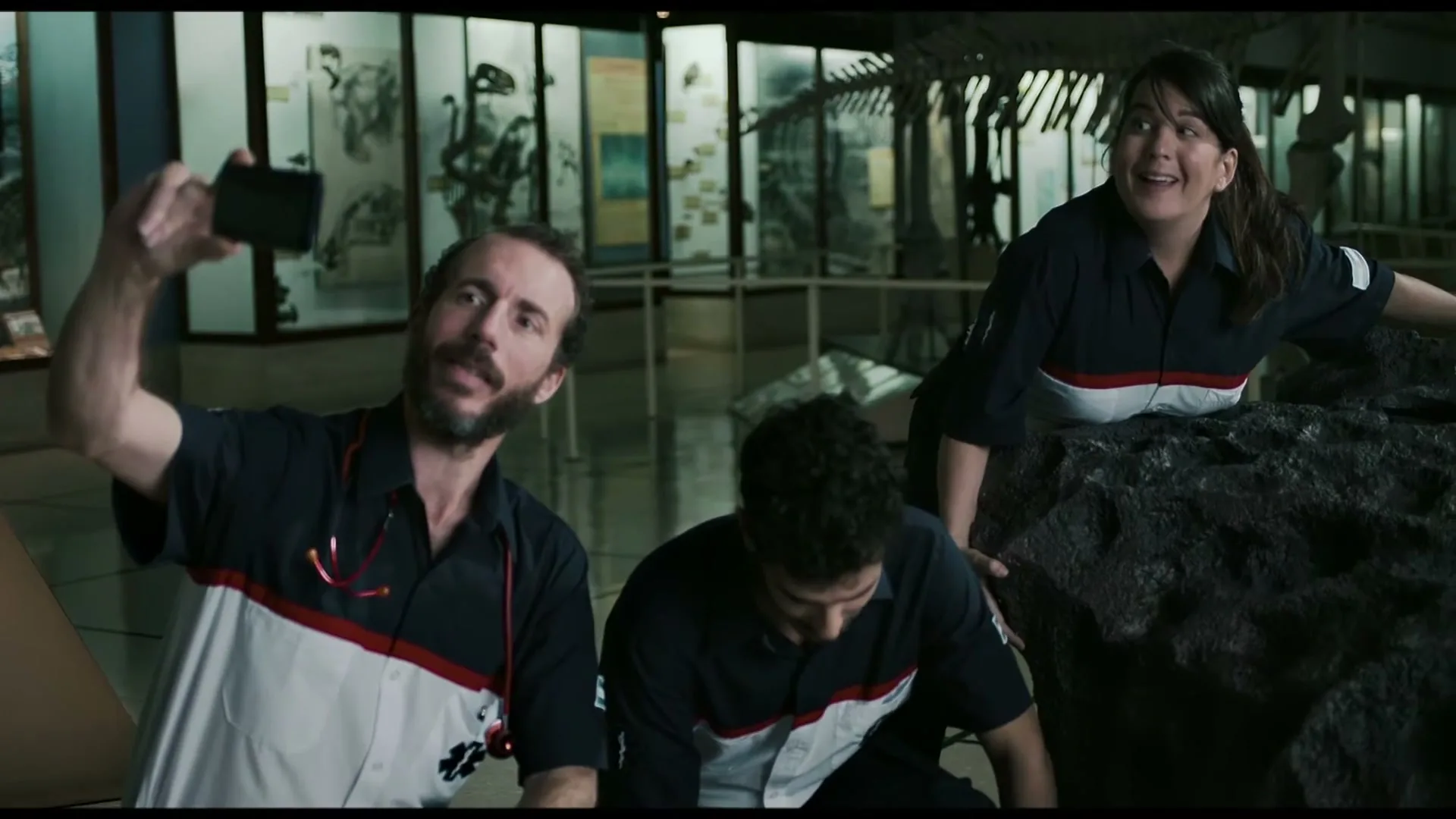 Three museum workers, two men and one woman, in blue and white uniforms, are posing for a selfie with a dinosaur skeleton in a dimly lit museum.