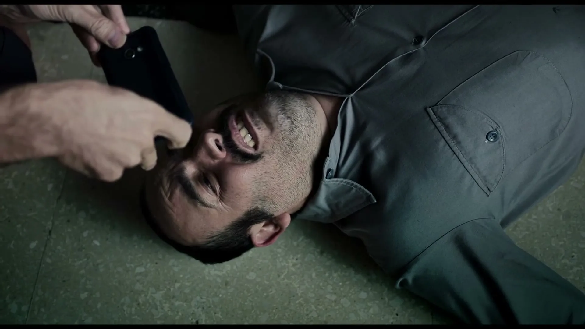 A man in his 30s, wearing a grey uniform, lies on the floor with a pained expression as another person takes a photo of him with a smartphone.