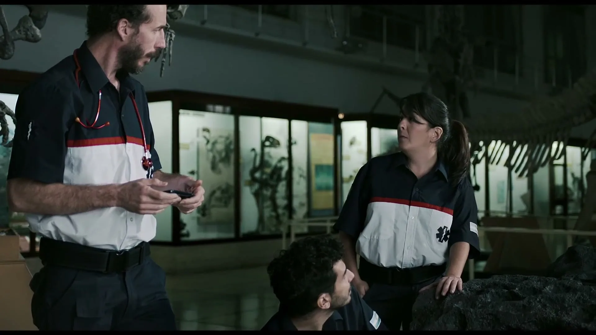 Three paramedics, two men and one woman, wearing blue, white, and red uniforms, are in a museum, looking at their phones and talking.