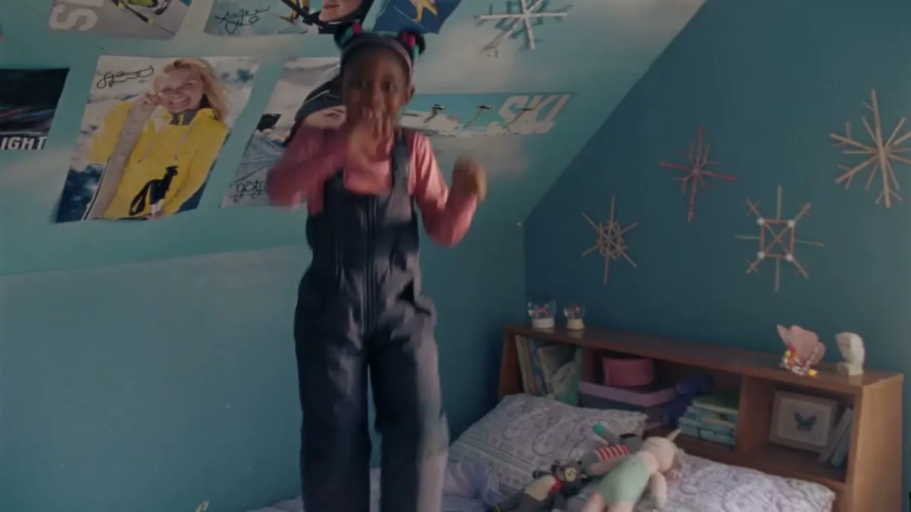 A young Black girl with pigtails and a pink shirt jumps on a bed in a blue bedroom decorated with ski posters.
