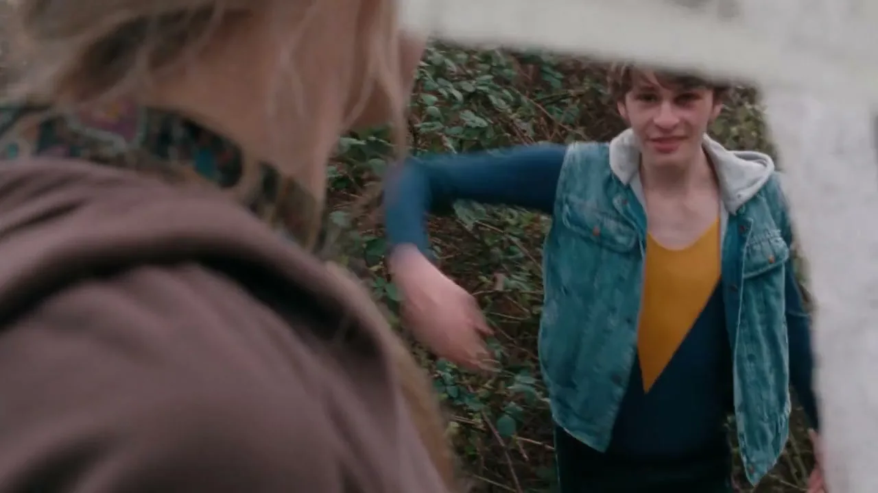 A young male dancer in a denim vest and yellow shirt runs and leaps gracefully towards the camera, then falls to the ground, seen from over the shoulder of another person outdoors.