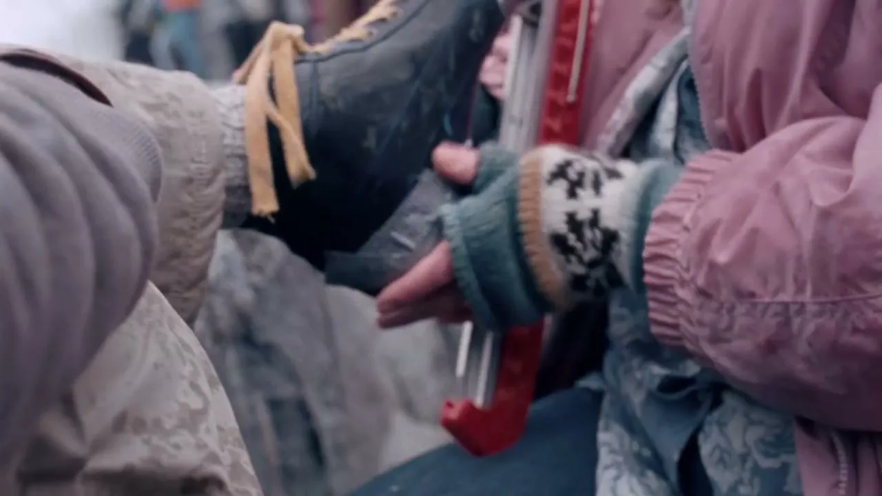 A person in a pink jacket and patterned mittens helps another person put on an ice skate, with other people visible in the blurry background.