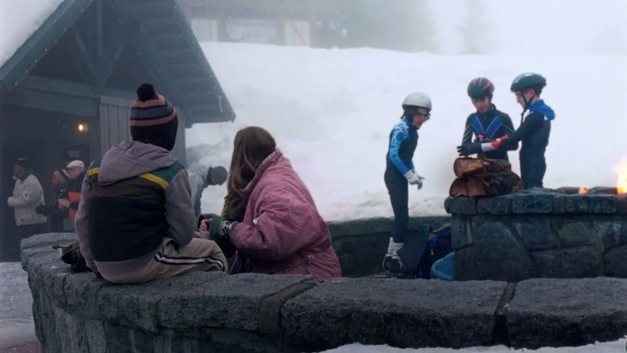 A group of people, including children in ski gear, gather around a stone fire pit in a snowy mountain resort, with a building in the background.