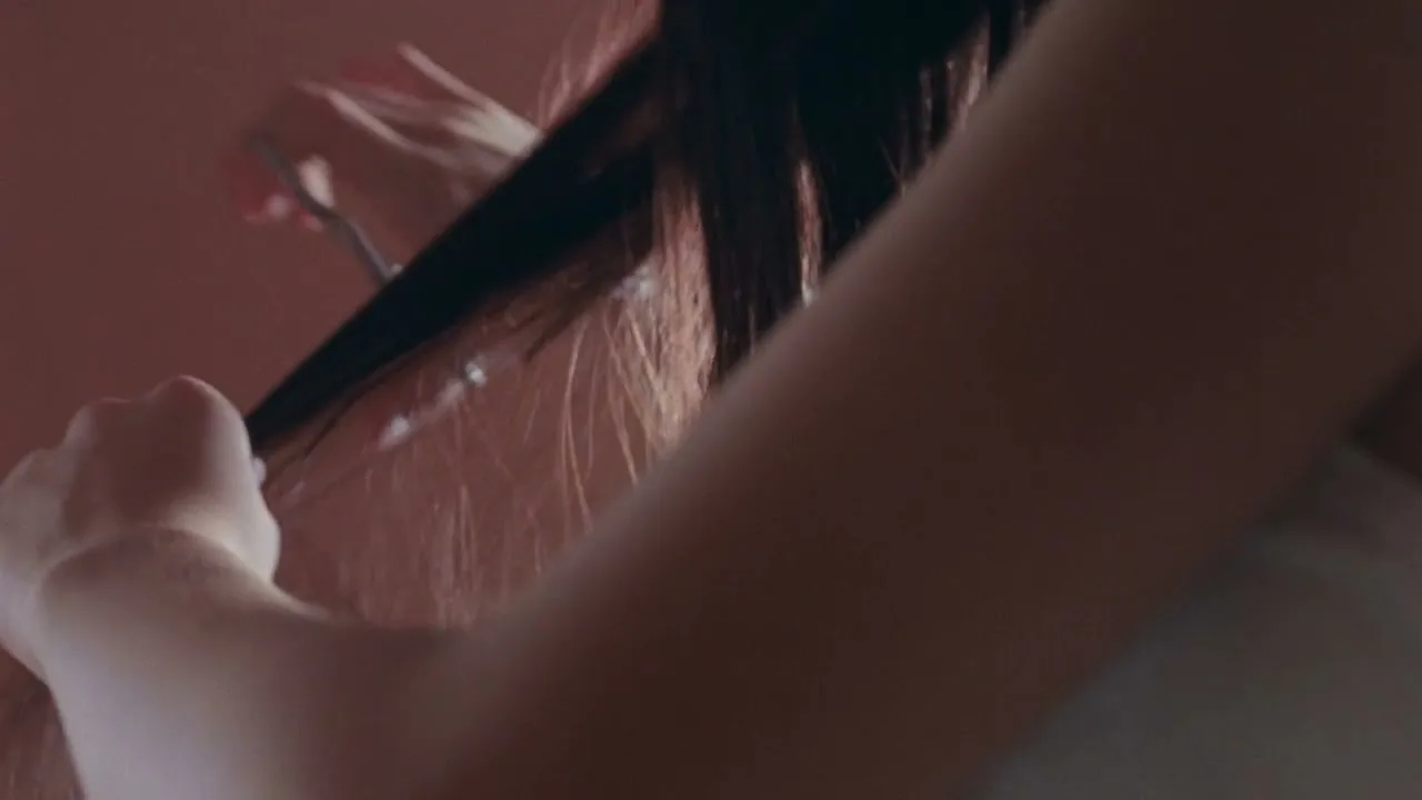A person with long dark hair is getting their hair cut by another person, visible only by their arm and hand holding scissors, in an interior setting.