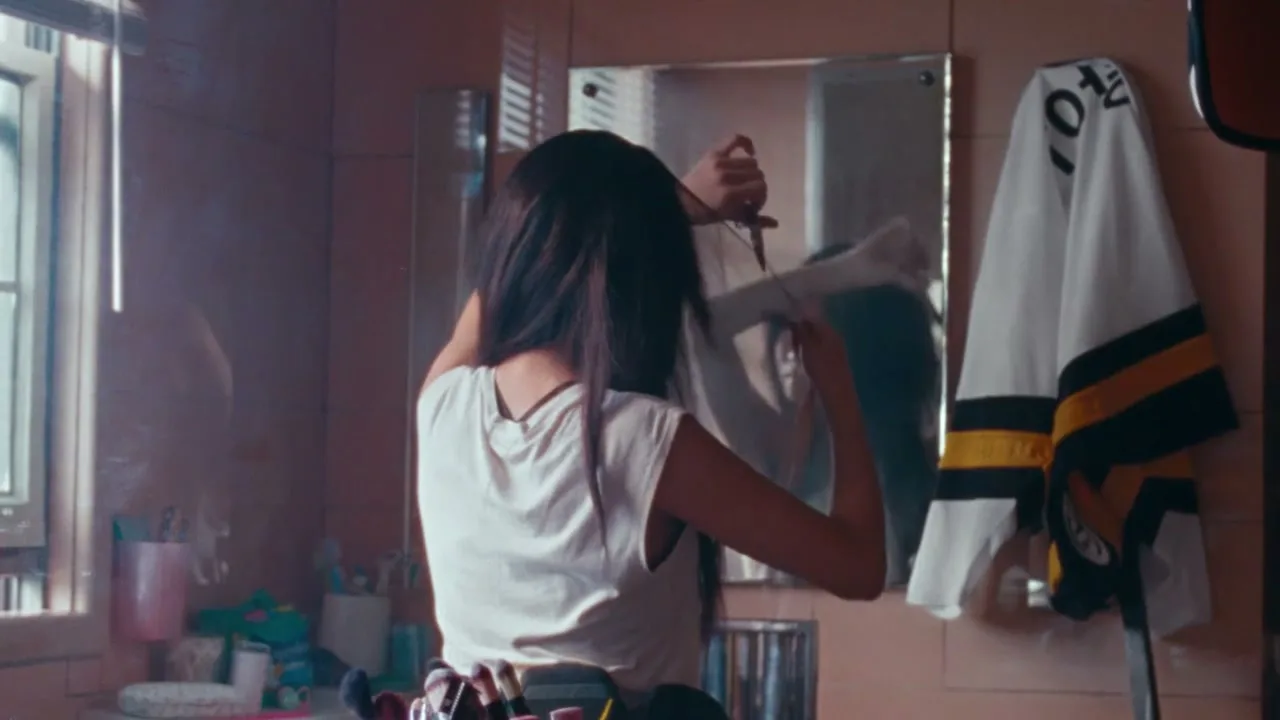 A young woman with long dark hair, seen from behind, styles her hair in a bathroom mirror, with a hockey jersey hanging nearby.
