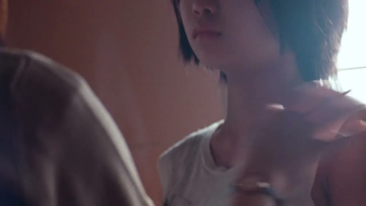 A young Asian woman with short dark hair looks on as an unseen person's arm reaches out to touch her shoulder in a warm, dimly lit room.