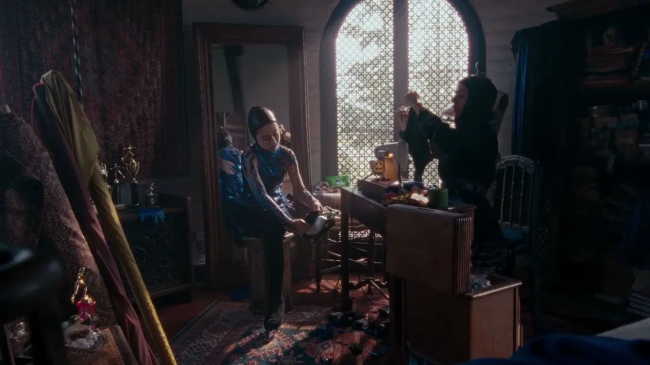 Two women, one in a blue sequined outfit and the other in black, are working on clothing in a cluttered, dimly lit room with a large arched window.