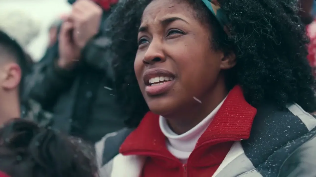 A young Black woman with curly hair, wearing a red and white winter jacket, looks up with a worried expression as snow falls around her in an outdoor stadium.