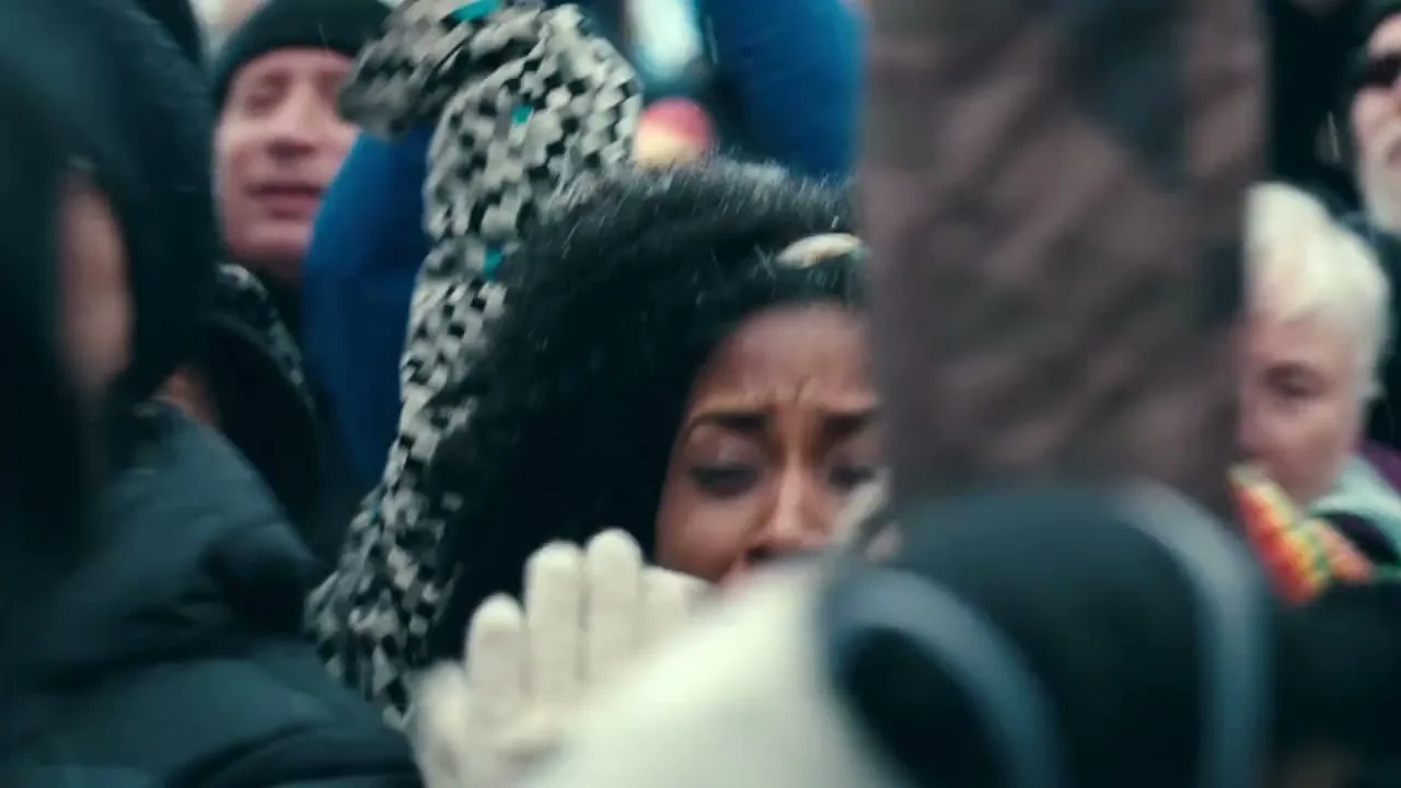 A diverse crowd of people, including a young Black woman with curly hair, are seen cheering and smiling at an outdoor event.