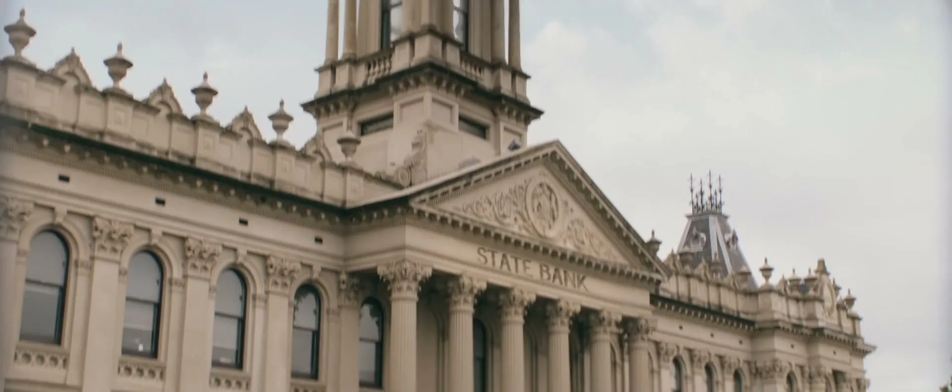 A clock tower and two helicopters are seen in the sky, then the camera pans down to reveal a bank building and people running from it, with police cars and officers on the street.