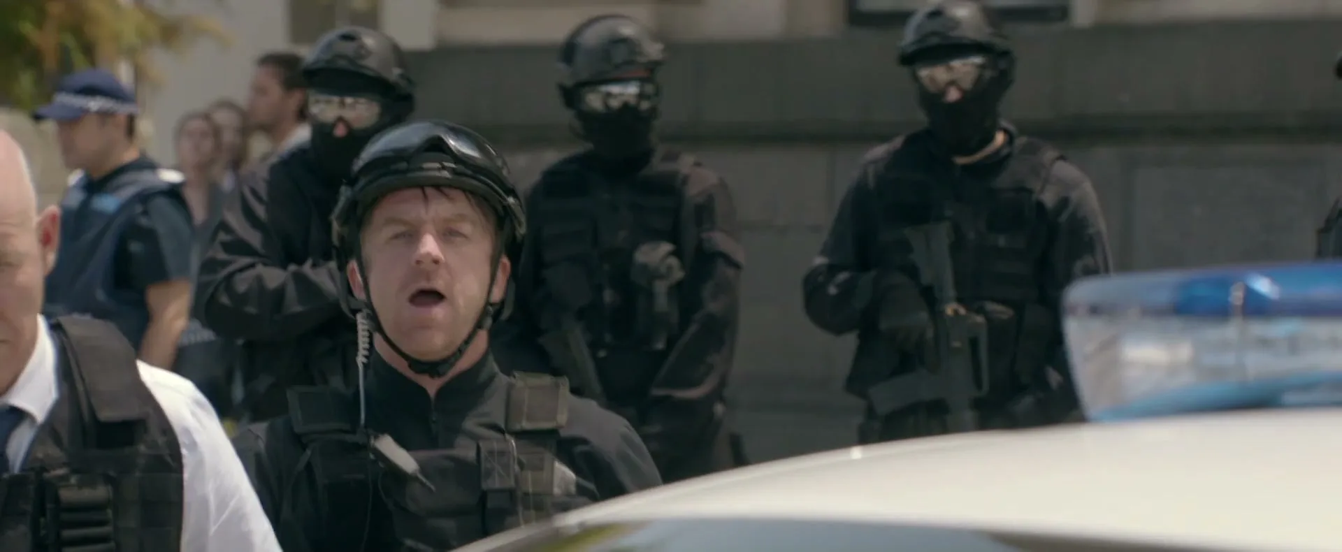 A male police officer in tactical gear looks up from behind a police car, while other officers stand in the background.