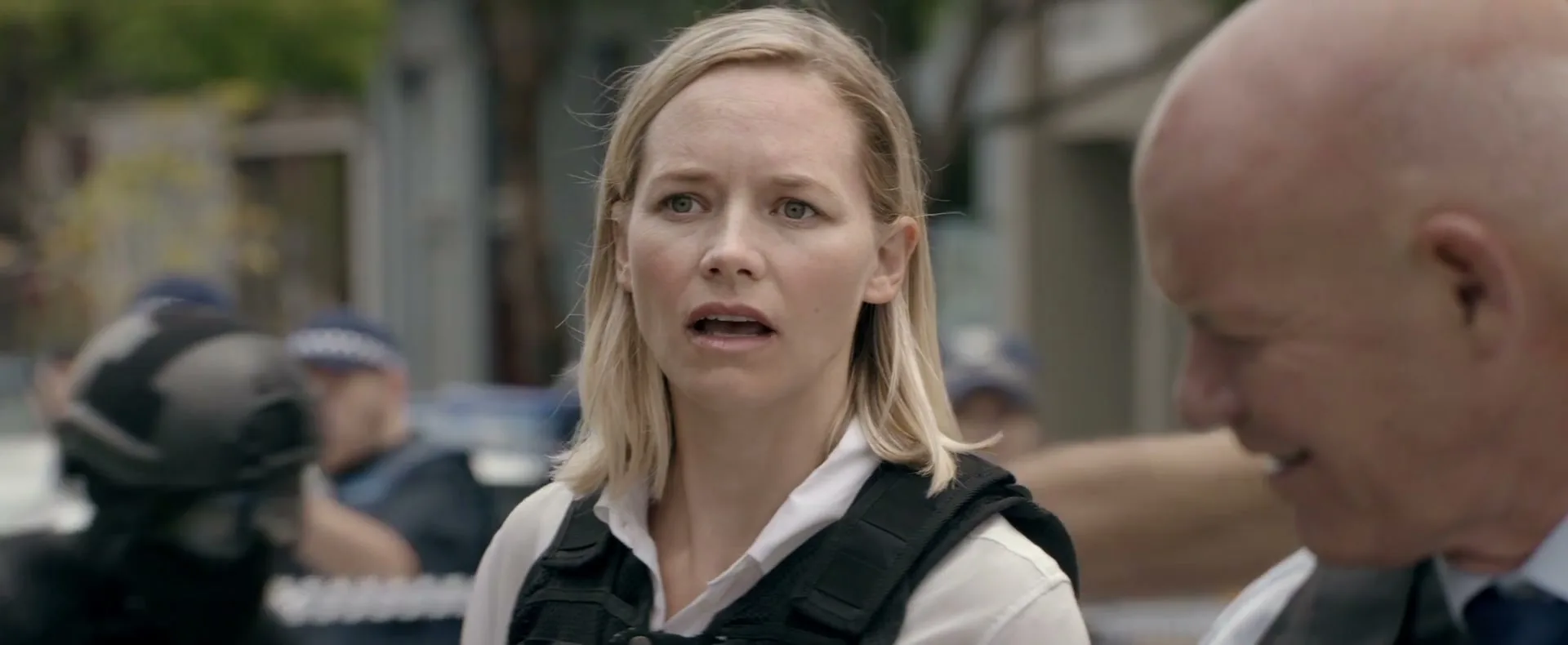 A blonde woman in a white shirt and black vest looks concerned while a bald man in a suit smiles beside her at a crime scene.