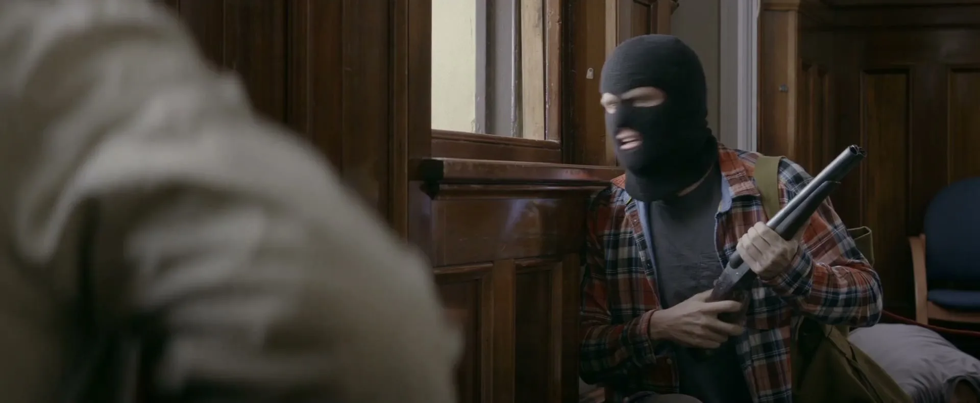 A man in a black balaclava and plaid shirt holds a shotgun, looking surprised as he peeks around a wooden doorframe in an old building.