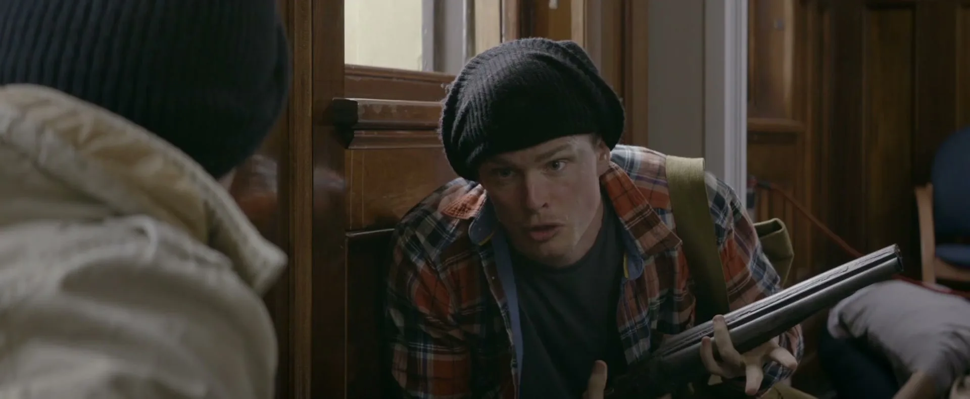 A man in a black beanie and plaid shirt holds a shotgun, looking surprised at another person in a beanie, indoors.