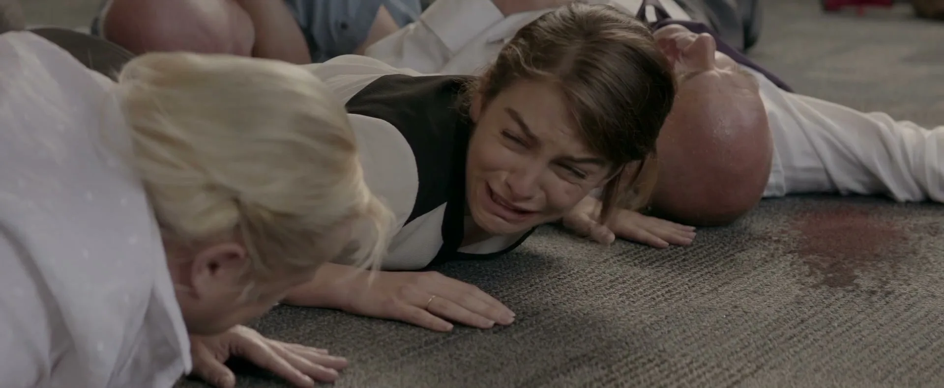 A woman in a white shirt and black vest lies on a carpeted floor, crying, surrounded by other people also lying on the floor, with a pool of blood nearby.