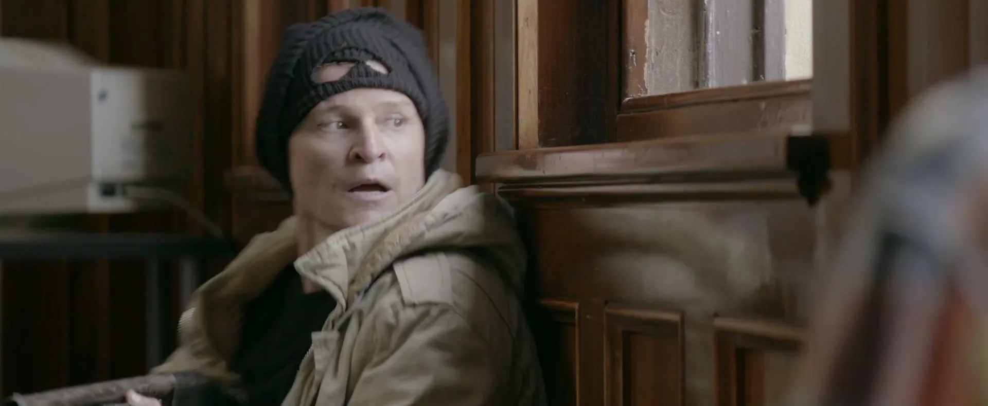 A man in a black beanie and a beige jacket looks surprised and scared, leaning against a wooden wall indoors.