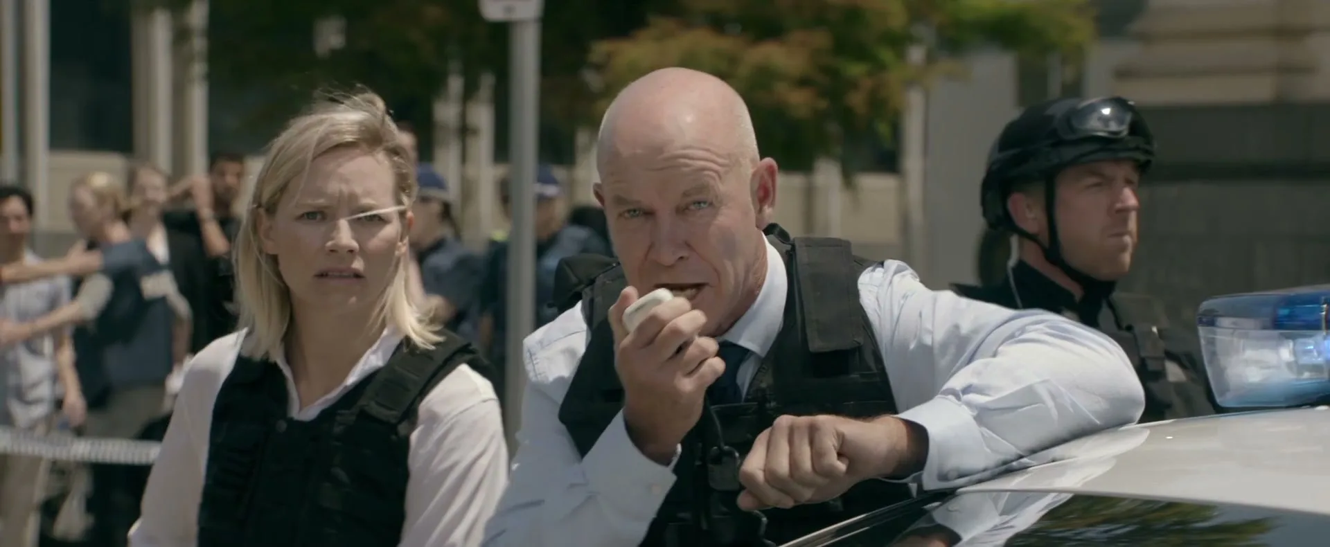 A male police officer in a bulletproof vest speaks into a radio while a female officer stands beside him, looking concerned, on a city street.