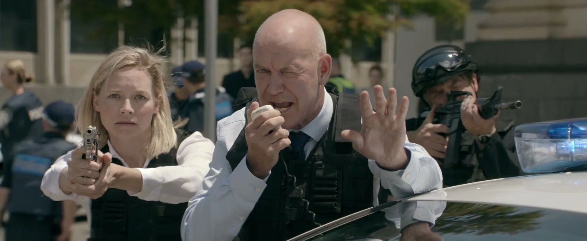 A bald man in a tactical vest speaks into a radio, holding up a hand to stop, while a blonde woman next to him aims a pistol, and another man aims a rifle behind a police car.