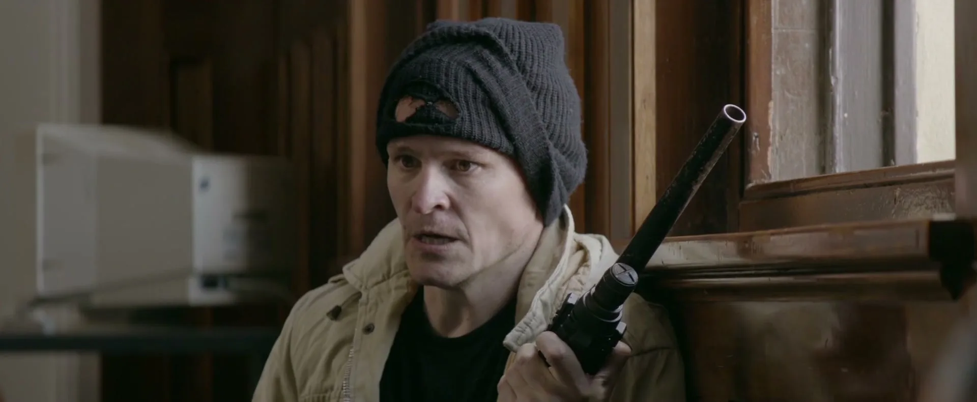 A man in a black beanie and beige jacket holds a shotgun, looking tense and pointing his finger in an old, wooden room.