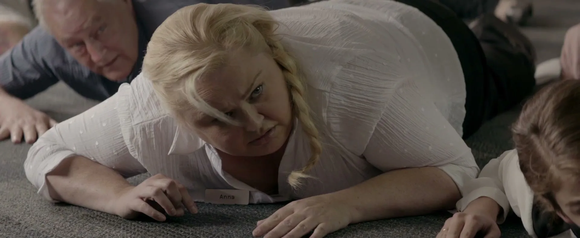 A blonde woman in a white shirt lies on the floor, looking intently at something off-screen, with an older man behind her.