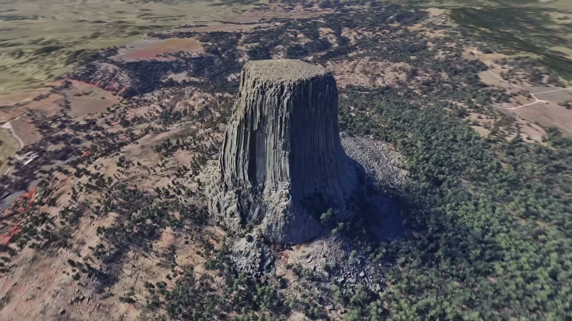 An aerial view shows the Devils Tower National Monument, a massive rock formation, surrounded by trees and a vast landscape under a clear sky.