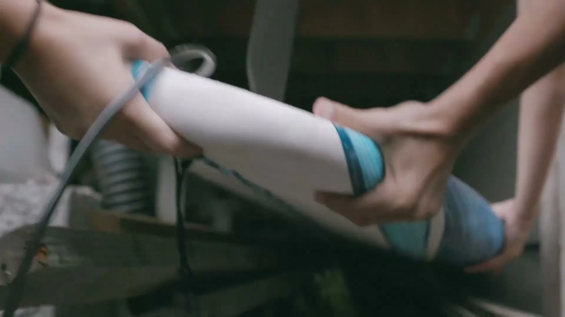 Two people's hands are seen from a low angle, holding a surfboard and preparing it for use in an interior setting.