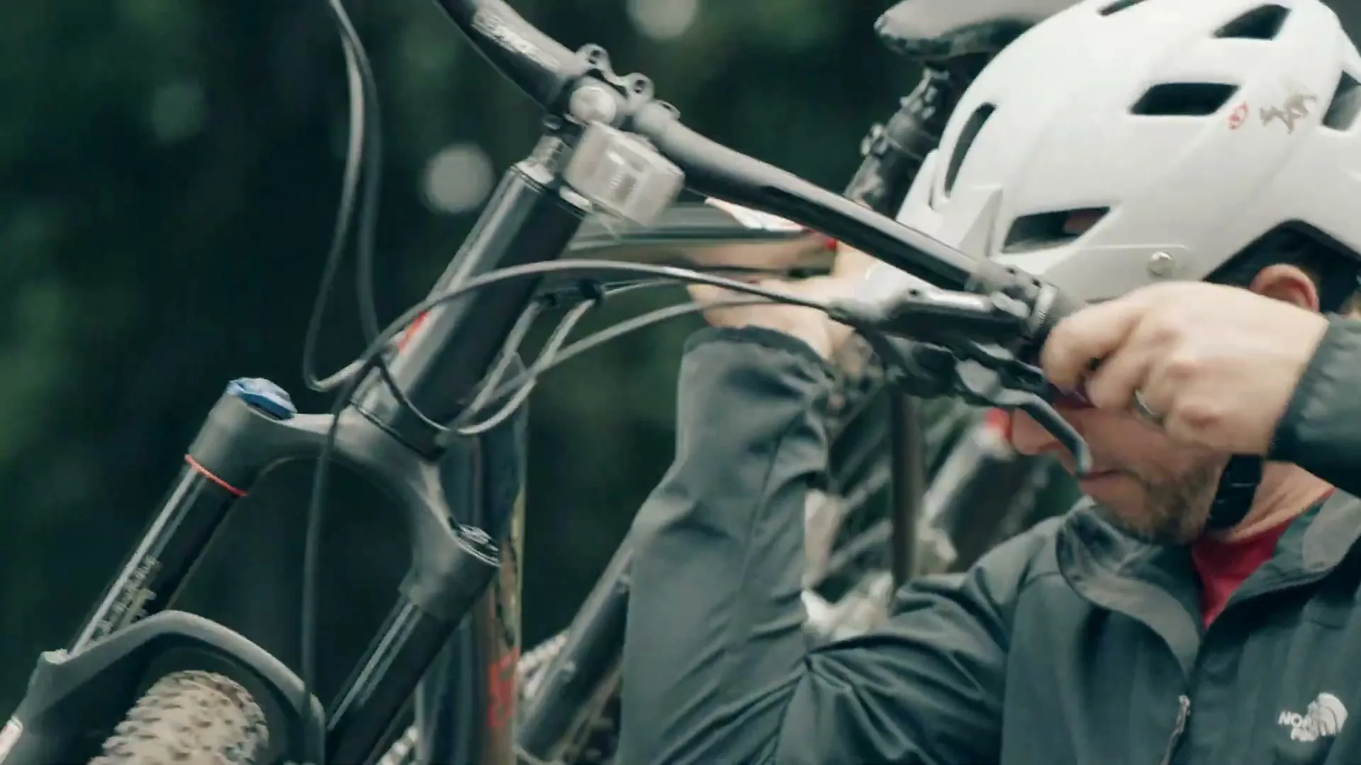 A man in a white helmet and black jacket is seen from the chest up, lifting a mountain bike off a car rack in an outdoor setting.