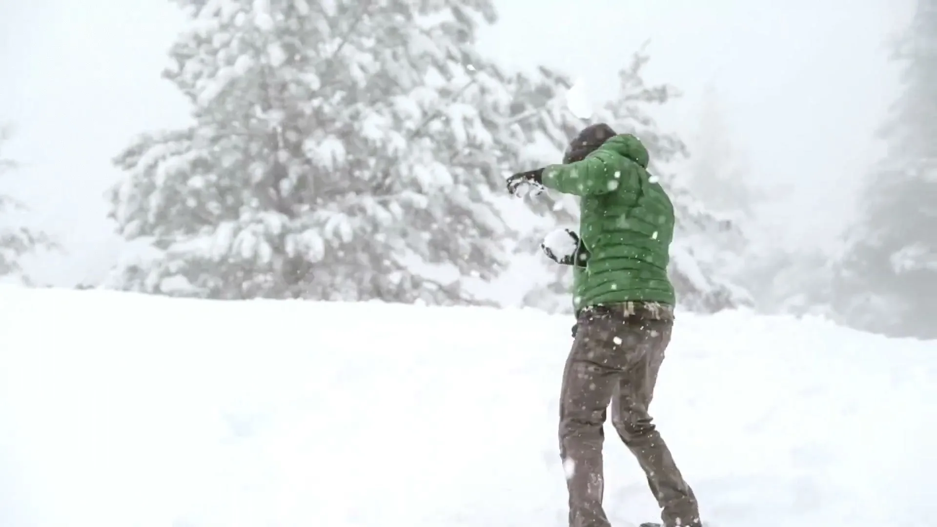 A man in a green jacket and brown pants is hit in the head by a snowball in a snowy forest.