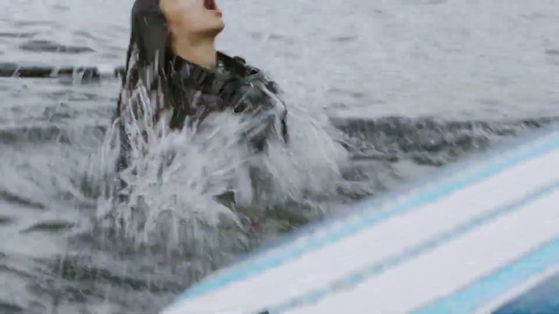 A young woman in a wetsuit emerges from the water next to a paddleboard, gasping, in a medium shot.