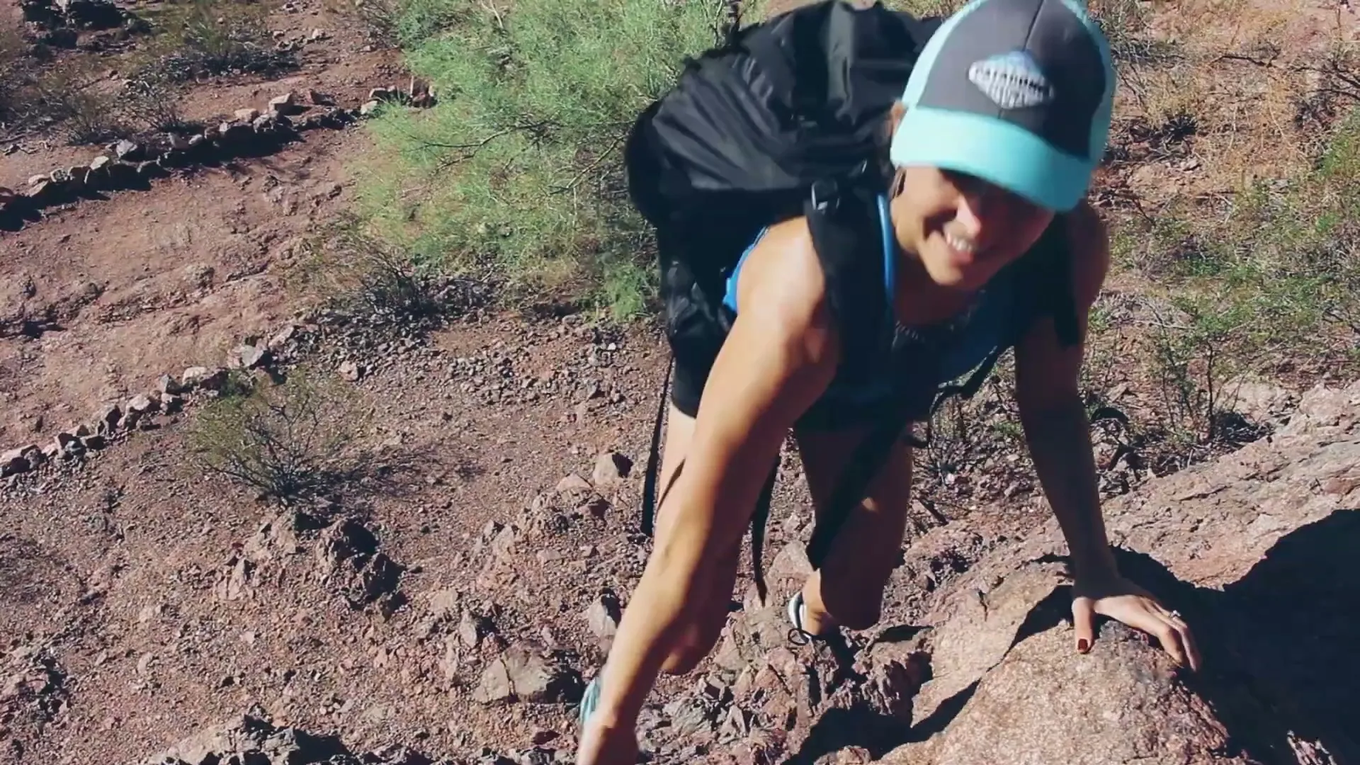 A young woman with a backpack and a cap climbs up a rocky, arid hill, smiling as she ascends.