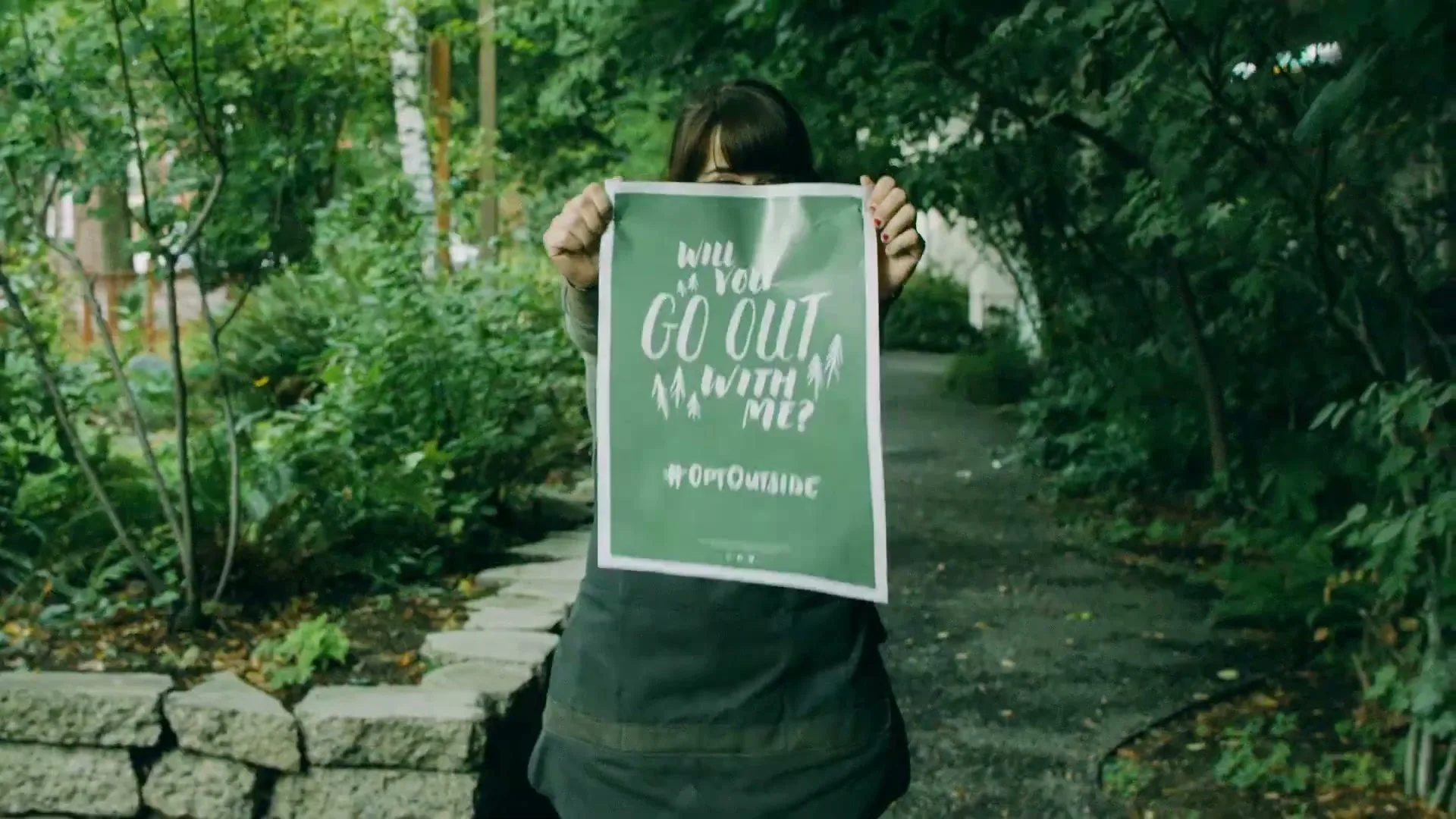 A young woman in glasses and an apron holds up a green sign that reads "Will you go out with me? #OptOutside" on a garden path.