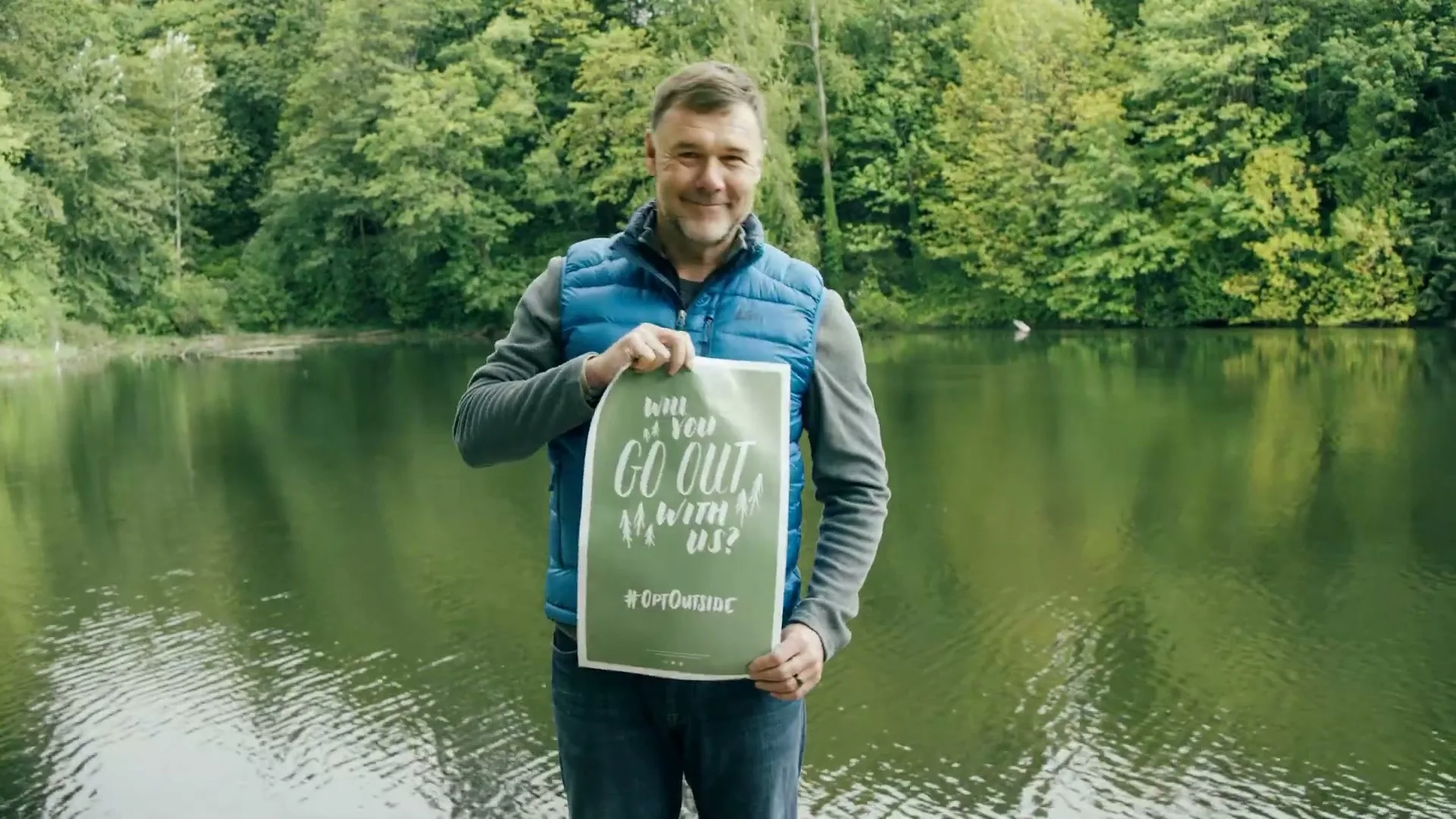 A man in his 40s, wearing a blue vest and jeans, holds a green sign that says "Will you go out with us? #OptOutside" by a lake surrounded by trees.