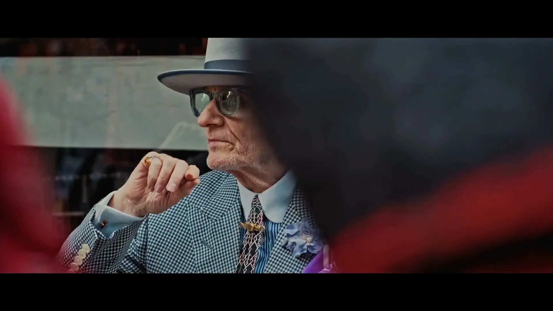 A stylish older man in a hat and glasses looks to the left, partially obscured by a red foreground element.