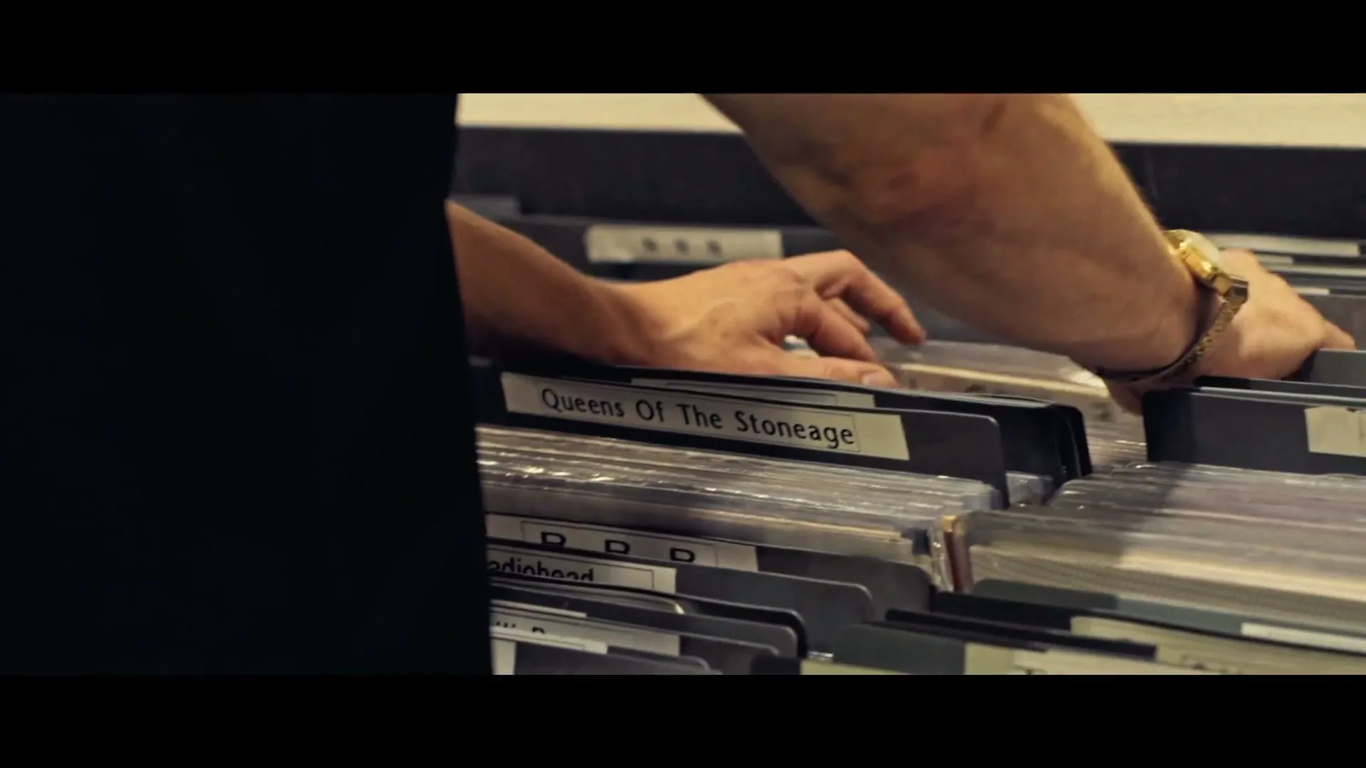 A person with a gold watch on their wrist is seen from the chest up, flipping through vinyl records in a record store.
